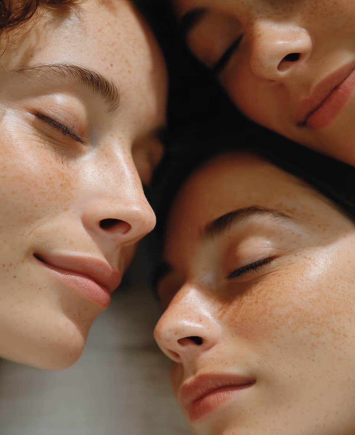 Close-up of three young women with closed eyes and relaxed expressions, showing natural skin and freckles.